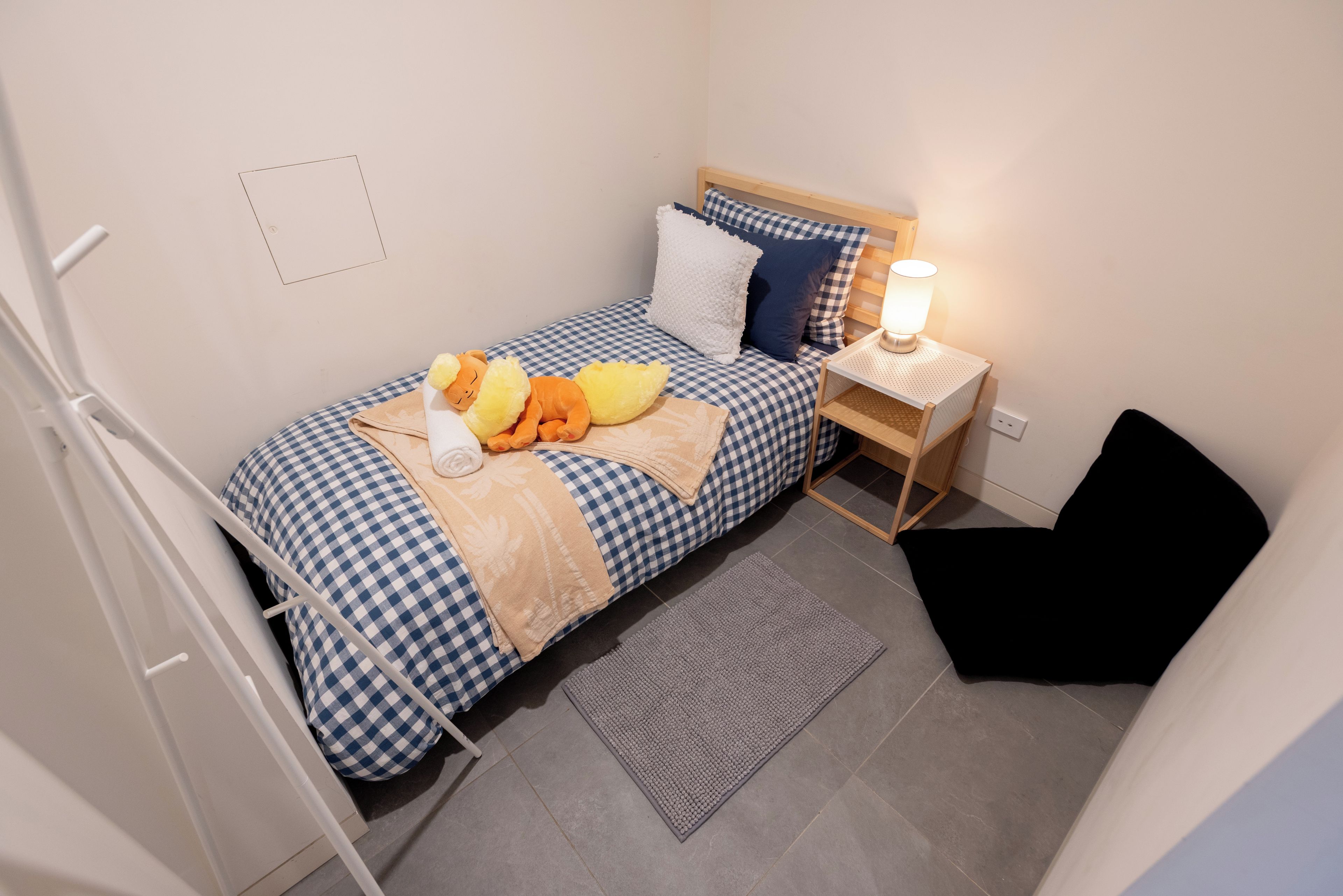 A single bed with a checkered bedspread, a bedside table with a lamp, a small rug, and a black chair.
