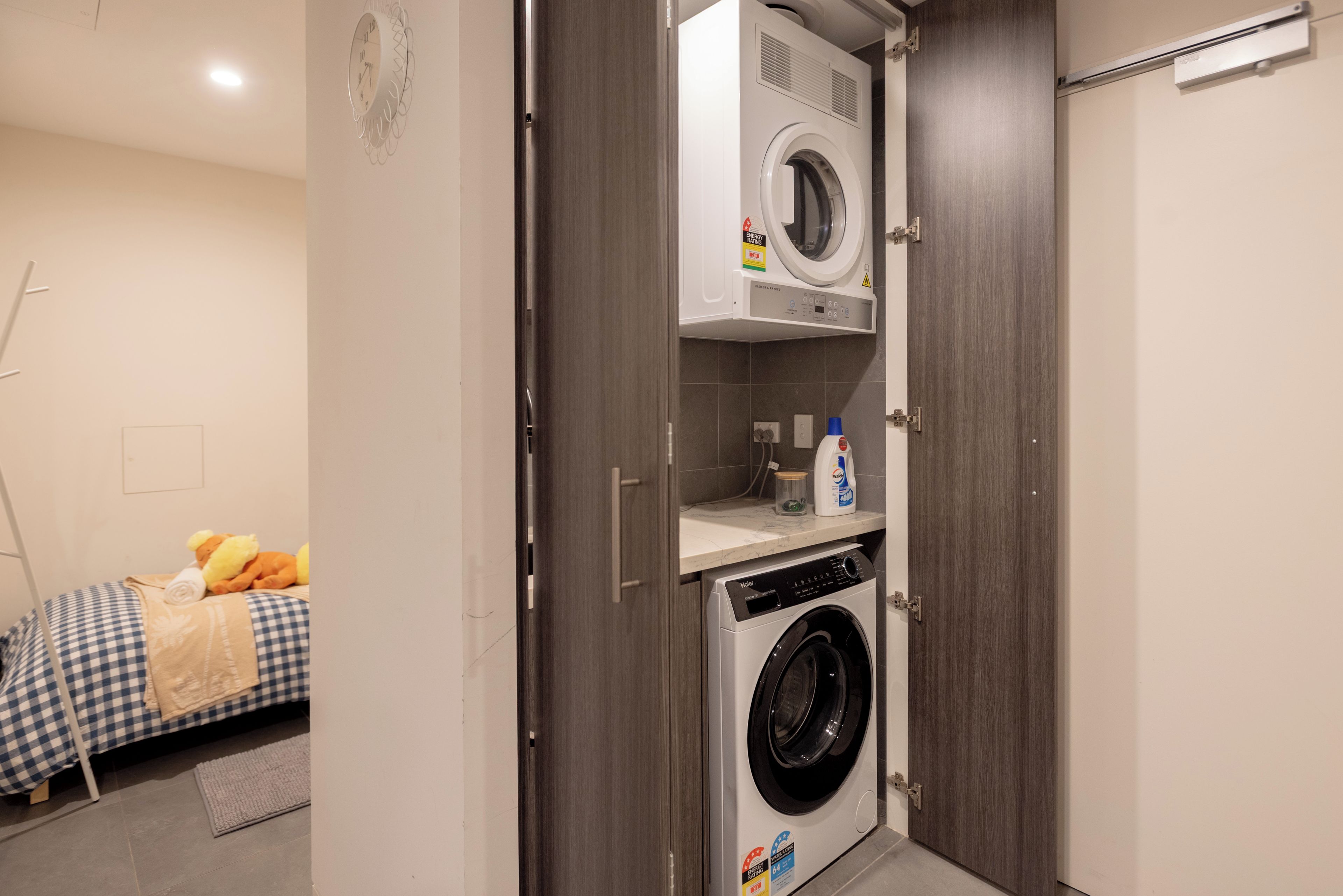 A compact laundry room with a front-loading washer and dryer, a small shelf with cleaning supplies, and a bed with a checkered bedspread in the background.