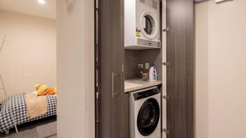 A compact laundry room with a front-loading washer and dryer, a small shelf with cleaning supplies, and a bed with a checkered bedspread in the background.