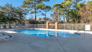 Piscina com cerca branca, cadeiras de descanso e árvores ao fundo.