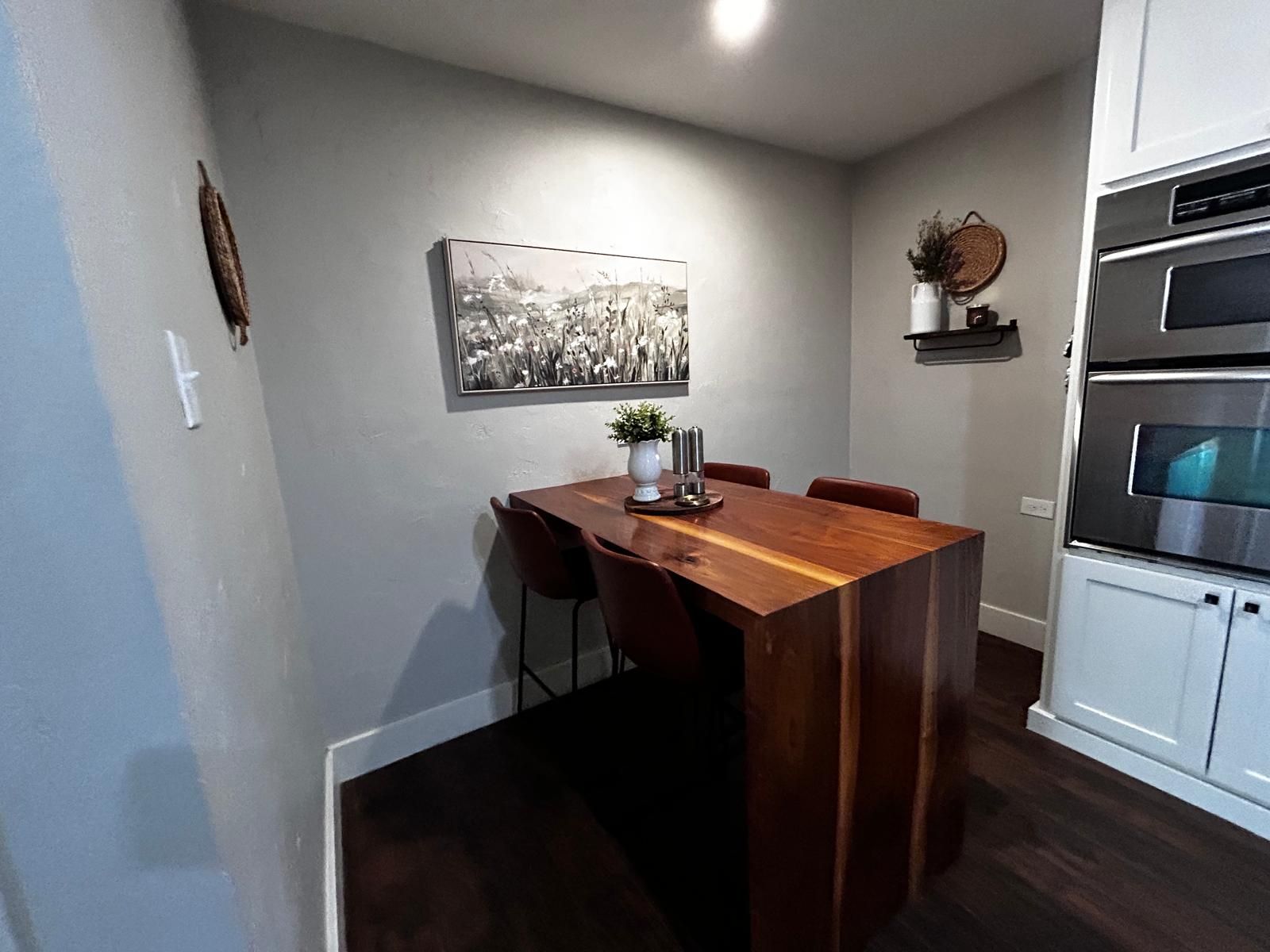 A compact kitchen with a wooden dining table, chairs, a hanging black and white photograph, and built-in ovens.