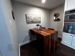 A compact kitchen with a wooden dining table, chairs, a hanging black and white photograph, and built-in ovens.