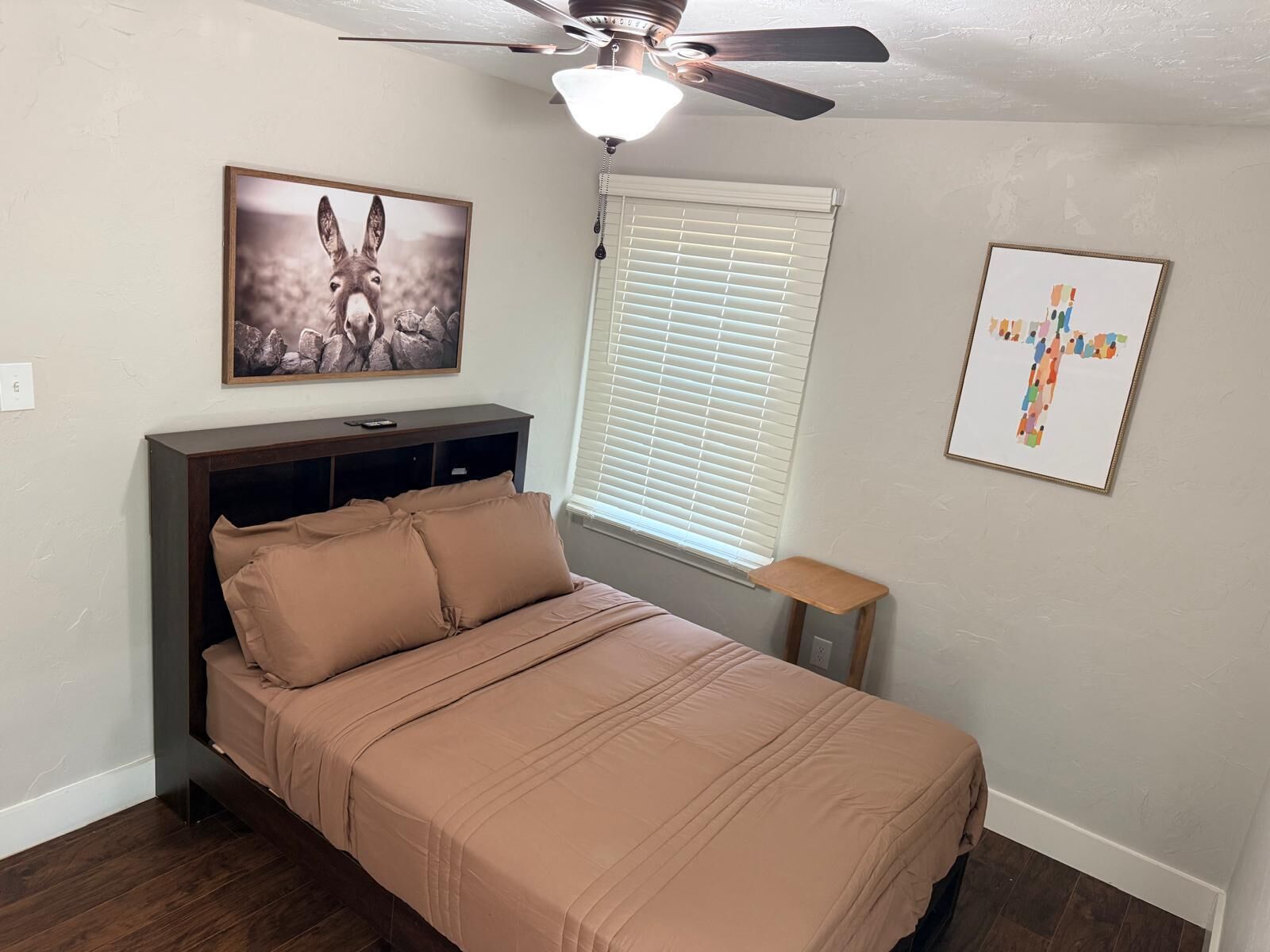 Une chambre à coucher avec un lit, un ventilateur de plafond, une fenêtre avec des stores et deux tableaux encadrés accrochés au mur.