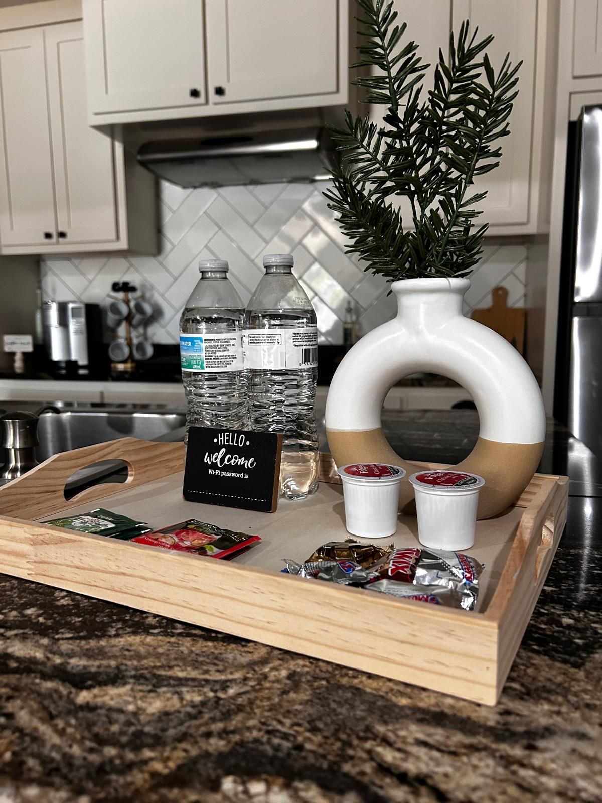 A tray with water bottles, a sign, snacks, and a decorative plant in a kitchen.