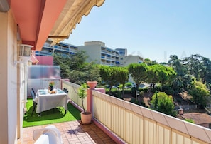 A balcony with a table set for dining, a potted plant, and a view of a residential area with trees and buildings. - Le Cappatti, 2 bedroom apartment (Nice)