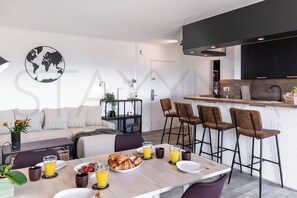 A modern kitchen with a bar area, a dining table set for breakfast, and a living area with a sofa and a world map decoration. - Le Cappatti, 2 bedroom apartment (Nice)