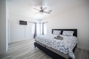 Ein Schlafzimmer mit einem Bett, einem Deckenventilator, einem an der Wand montierten Fernseher und einem Fenster mit Vorhängen.