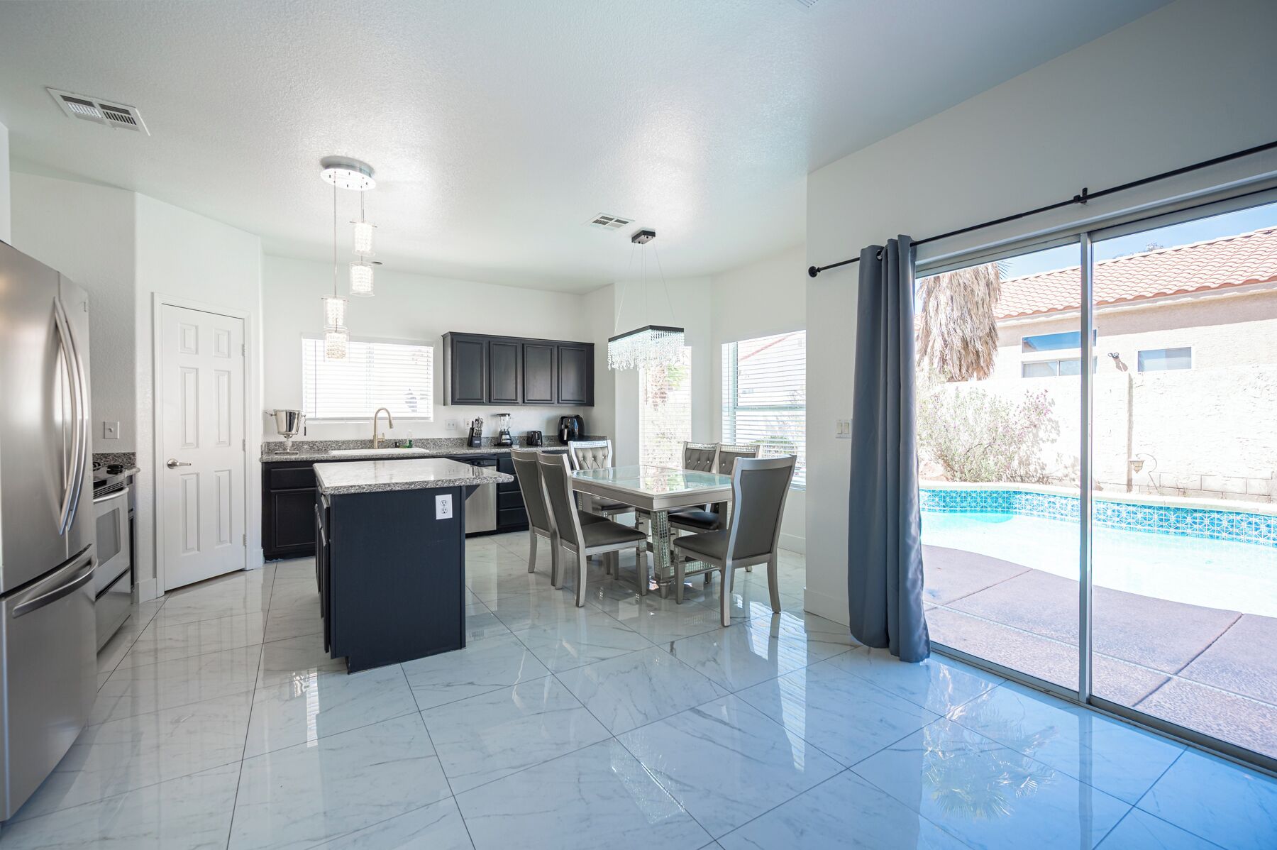 A modern kitchen with a central island, a dining area with a glass table, and a view of a swimming pool through large sliding doors.