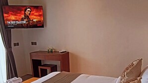 A hotel room with a bed, a desk, a television displaying a movie, and a window with curtains. - Cozy 1-bedroom hotel suite in enchanting Ubud
with AC (Gianyar)