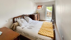 A bedroom with a bed, a wooden nightstand, a small table, and a window with curtains. - Lovely home in Châteauneuf-Villevieil (Châteauneuf-Villevieil)
