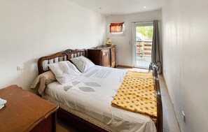 A bedroom with a bed, a wooden nightstand, a small table, and a window with curtains. - Lovely home in Châteauneuf-Villevieil (Châteauneuf-Villevieil)