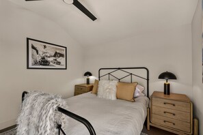Une chambre à coucher avec un lit, deux tables de chevet, un ventilateur de plafond, un tableau encadré au mur et une chaise recouverte d’un plaid en fausse fourrure.
