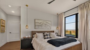 A bedroom with a large bed, bedside tables, a nightstand, a window with curtains, and artwork on the wall. - Teton Sunset 3BR luxury home sleeps 12 with private hot tub & unbeatable Teton mtn views. (Driggs)