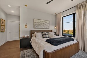 A bedroom with a large bed, bedside tables, a nightstand, a window with curtains, and artwork on the wall. - Teton Sunset 3BR luxury home sleeps 12 with private hot tub & unbeatable Teton mtn views. (Driggs)
