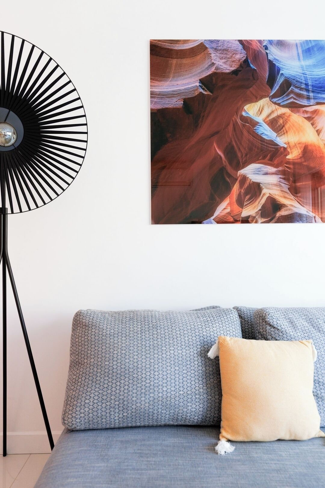 Een kamer met een grijze bank, een zwarte vloerlamp en een wanddecoratie met abstracte kunst.