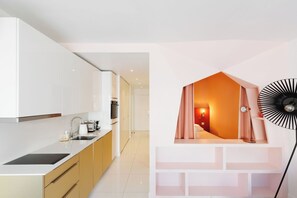 A modern kitchen with white cabinets, a sink, and a stove. There is a bedroom with a bed and a unique orange wall. A black fan is visible in the room. - Upstays Elegant Gray d'Albion 2min Palais (Cannes)