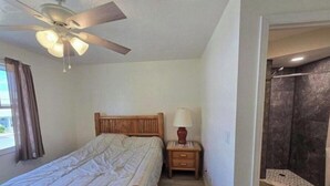 A bedroom with a bed, nightstand, lamp, ceiling fan, and a shower area.