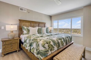 A bedroom with a large bed, a nightstand, a lamp, a bench, and a window with a view. - New listing! Updated oceanfront 2/2 with private balcony, heated pool, and garage parking (Ormond Beach)