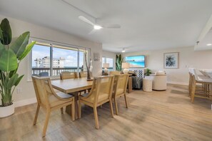 A modern dining area with a wooden table and chairs, a view of the city through large windows, and a ceiling fan. - New listing! Updated oceanfront 2/2 with private balcony, heated pool, and garage parking (Ormond Beach)