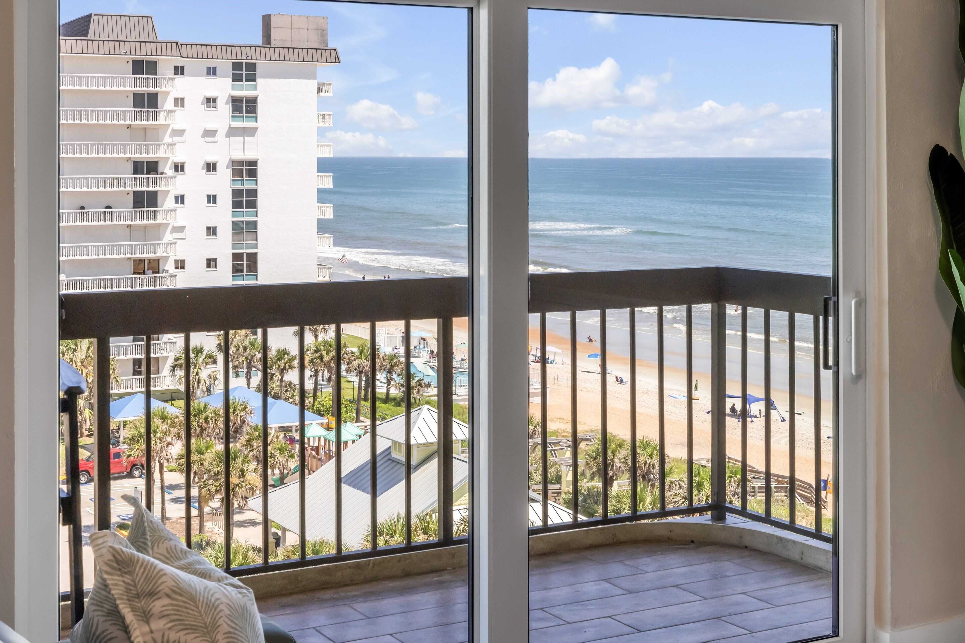 Ein Balkon mit Blick auf Strand und Meer, ein Gebäude im Hintergrund sowie eine Palme.