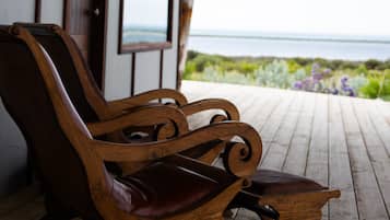 A wooden bench with a cushioned seat and backrest, positioned on a wooden deck with a view of the sea and greenery in the background.