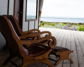 A wooden bench with a cushioned seat and backrest, positioned on a wooden deck with a view of the sea and greenery in the background.