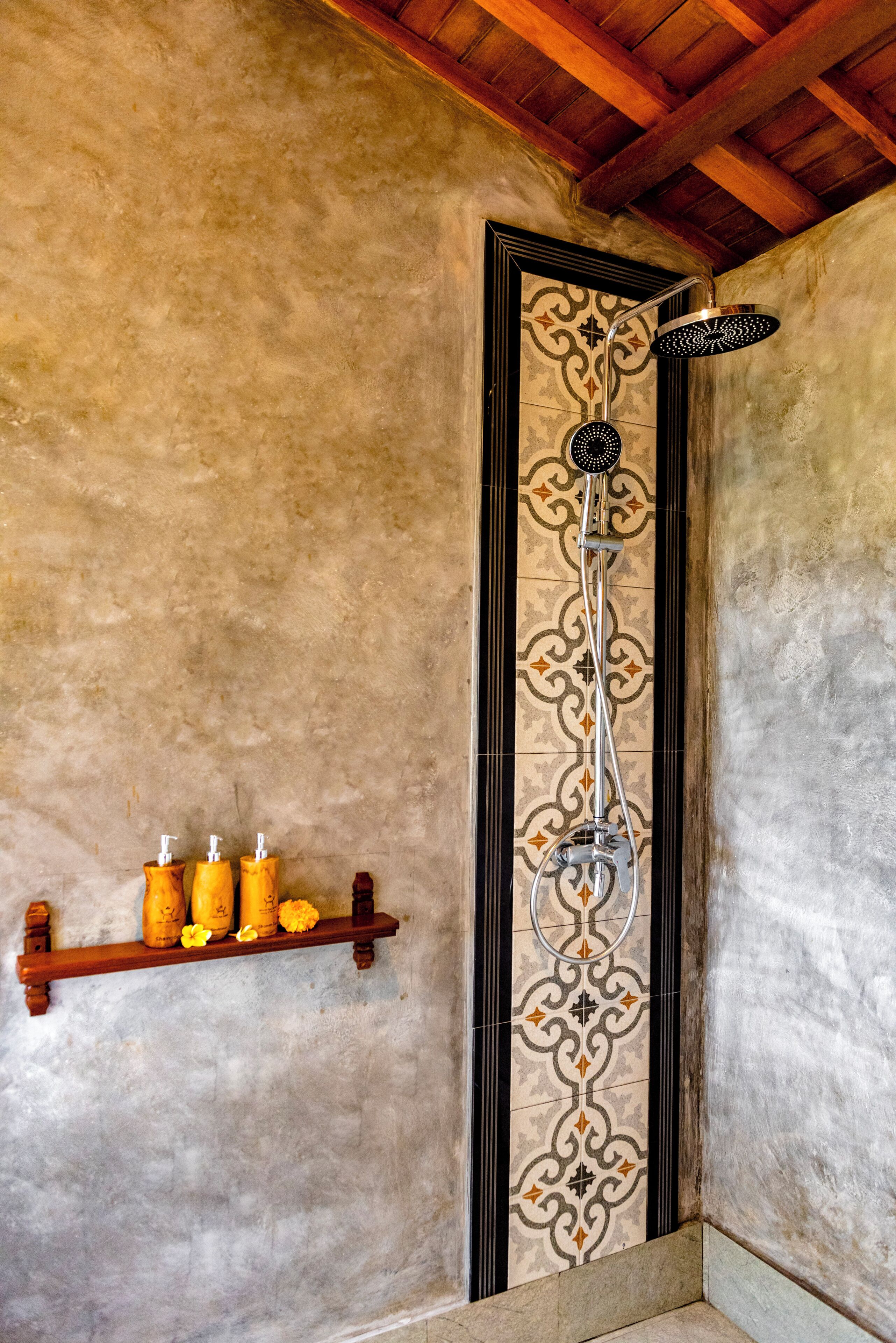 Una doccia con una parete rivestita di piastrelle decorate, uno scaffale in legno con articoli da toilette e un soffitto in legno.