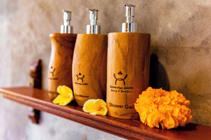 Tres botellas de madera etiquetadas como 'Shower Gel' con dosificador de bomba, sobre una estantería de madera junto a una flor amarilla.