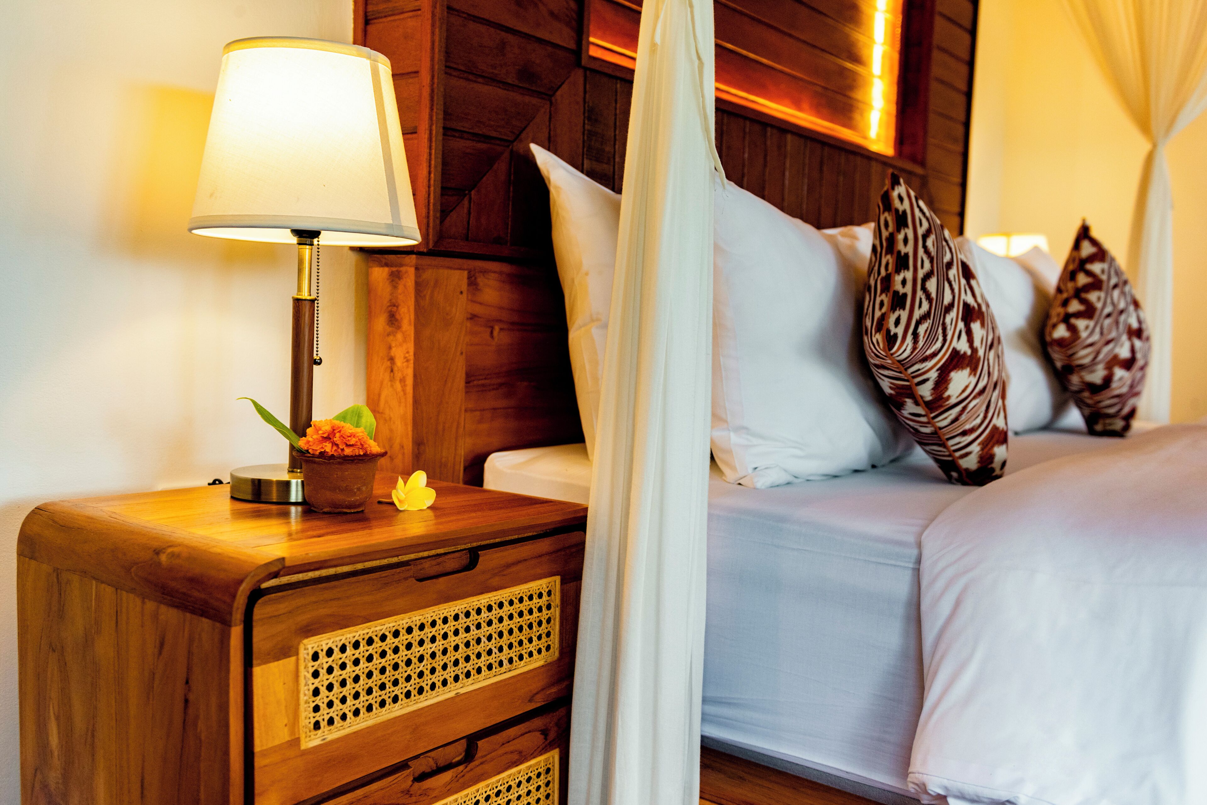 Una cama bien hecha con sábanas blancas, una mesita de noche de madera con una lámpara y almohadones decorativos.