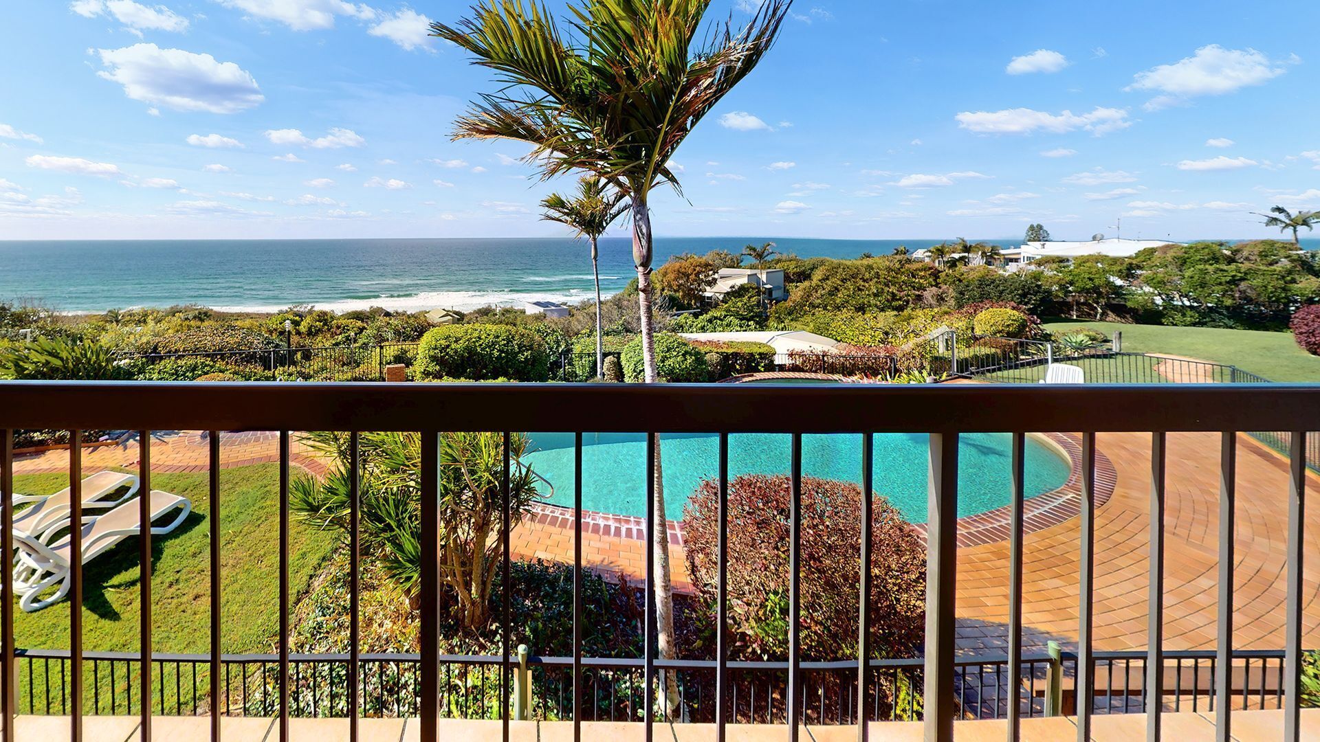A balcony with a view of the ocean, a swimming pool, and a landscaped garden.