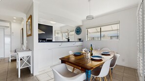 A modern dining area with a wooden table set for four, white chairs, and a kitchen in the background. - Sunseeker Lodge – Ocean View in Sunshine Beach (Sunshine Beach)