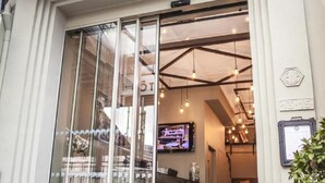 Entrada de hotel com porta de vidro, televisão montada na parede e teto com vigas aparentes e luminárias pendentes.