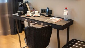 Quarto de hotel moderno com uma secretária, uma cadeira, uma televisão na parede, uma máquina de café, uma garrafa de água e uma revista.