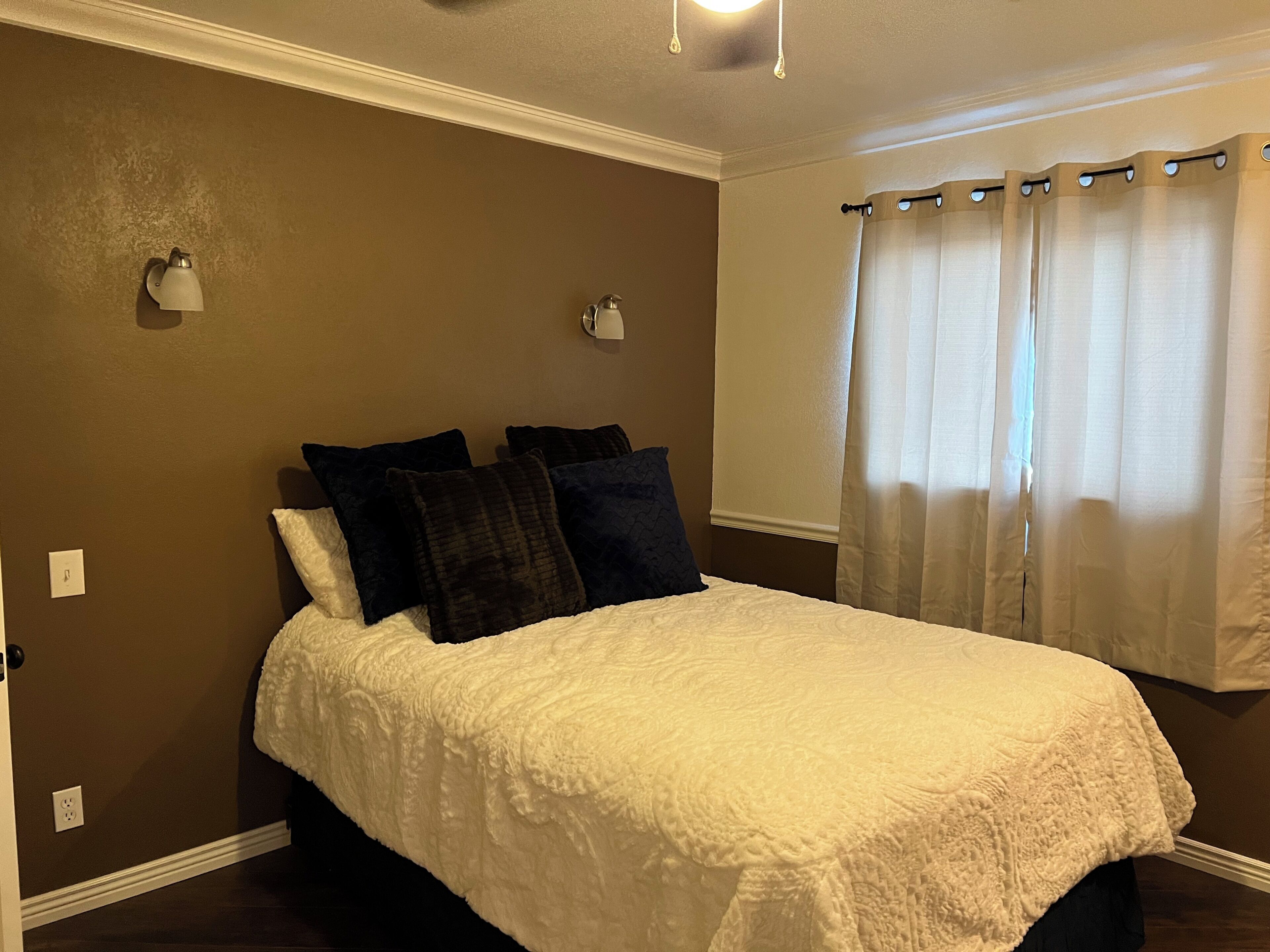 A bedroom with a bed, two wall-mounted lights, a window with curtains, and a wall switch.