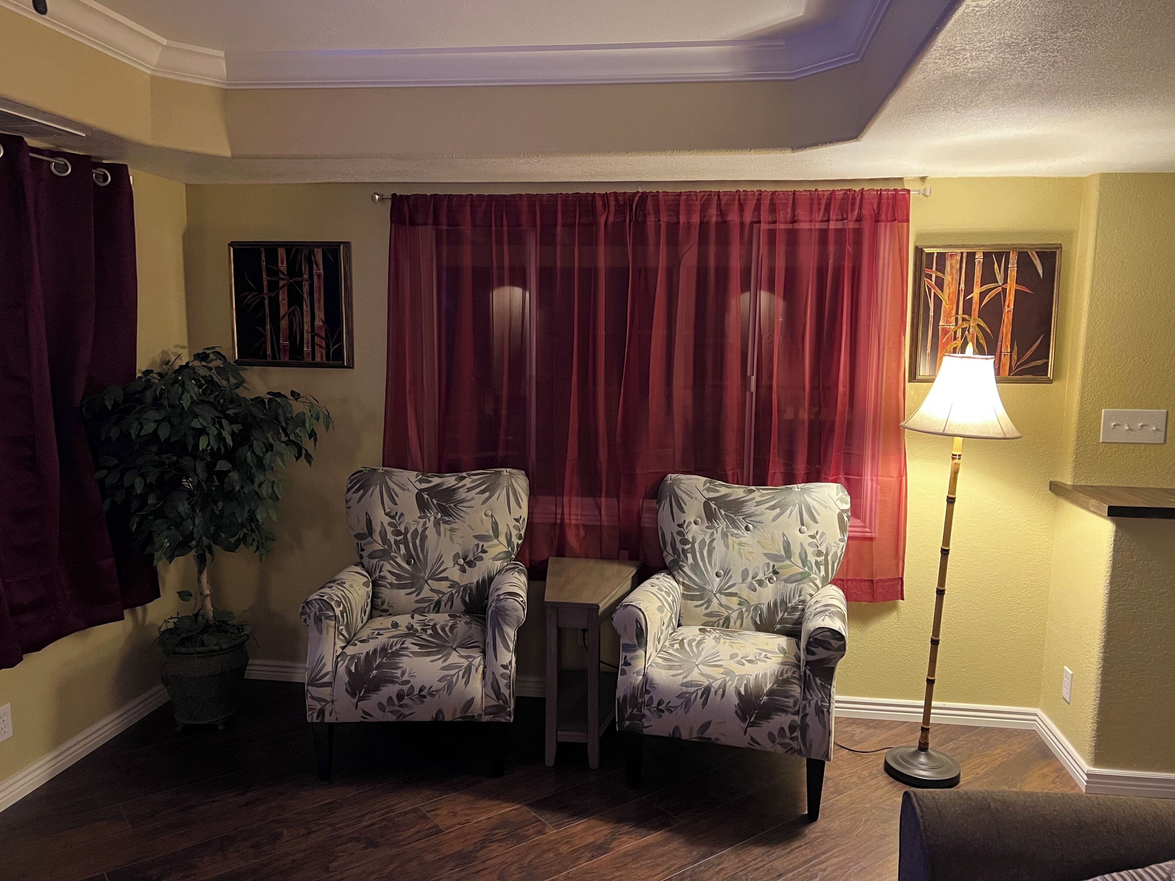 A living room with two patterned armchairs, a small table, a floor lamp, a potted plant, and a painting on the wall.