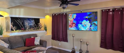 Una sala de estar con un sofá gris, un acuario, un televisor de pantalla plana y un ventilador de techo.