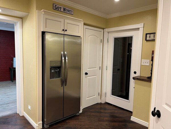 A kitchen with a stainless steel refrigerator, white cabinetry, and a door with a frosted glass panel. - The Bar House (Seligman)