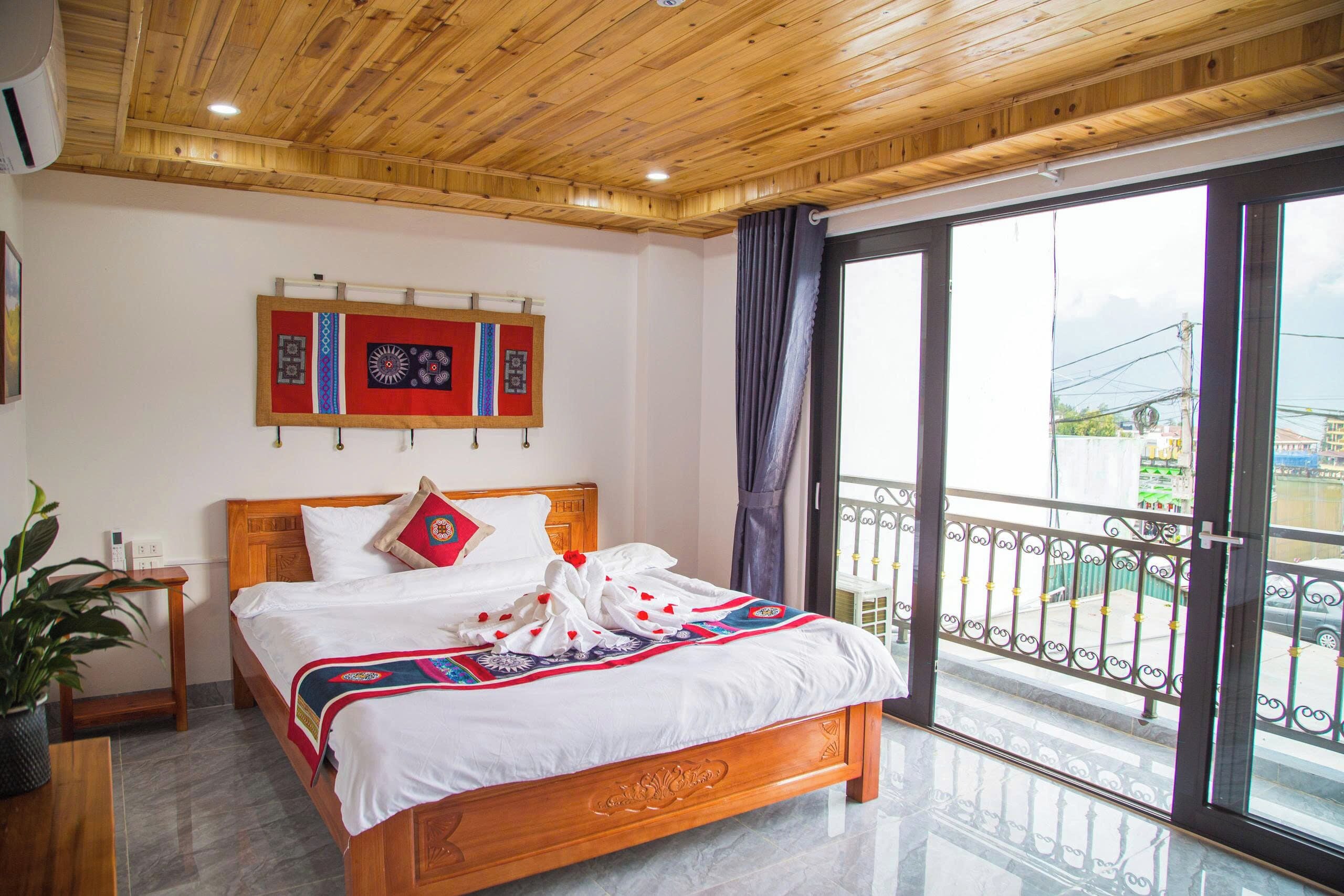 Une chambre d’hôtel avec un plafond en bois, un grand lit recouvert d’une couverture à motifs rouges et bleus, une table de chevet avec un téléphone, et une vue sur l’extérieur grâce à un balcon doté d’une rambarde en métal.
