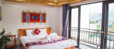 Habitación de hotel con techo de madera, cama grande con colcha de diseño rojo y azul, mesita de noche con teléfono y vista al exterior a través de un balcón con barandilla metálica.