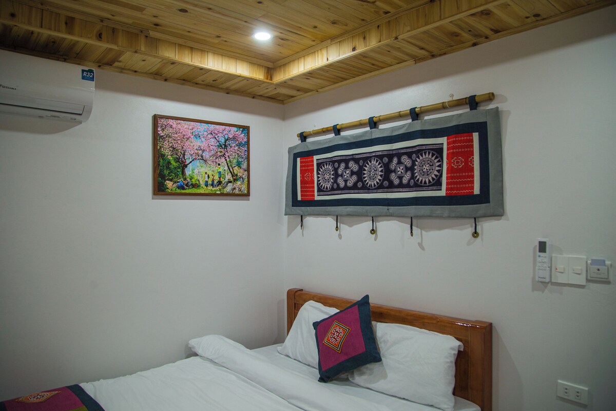 Un dormitorio con techo de madera, una cama con ropa blanca, una almohada colorida, un aire acondicionado de pared y un cuadro enmarcado.