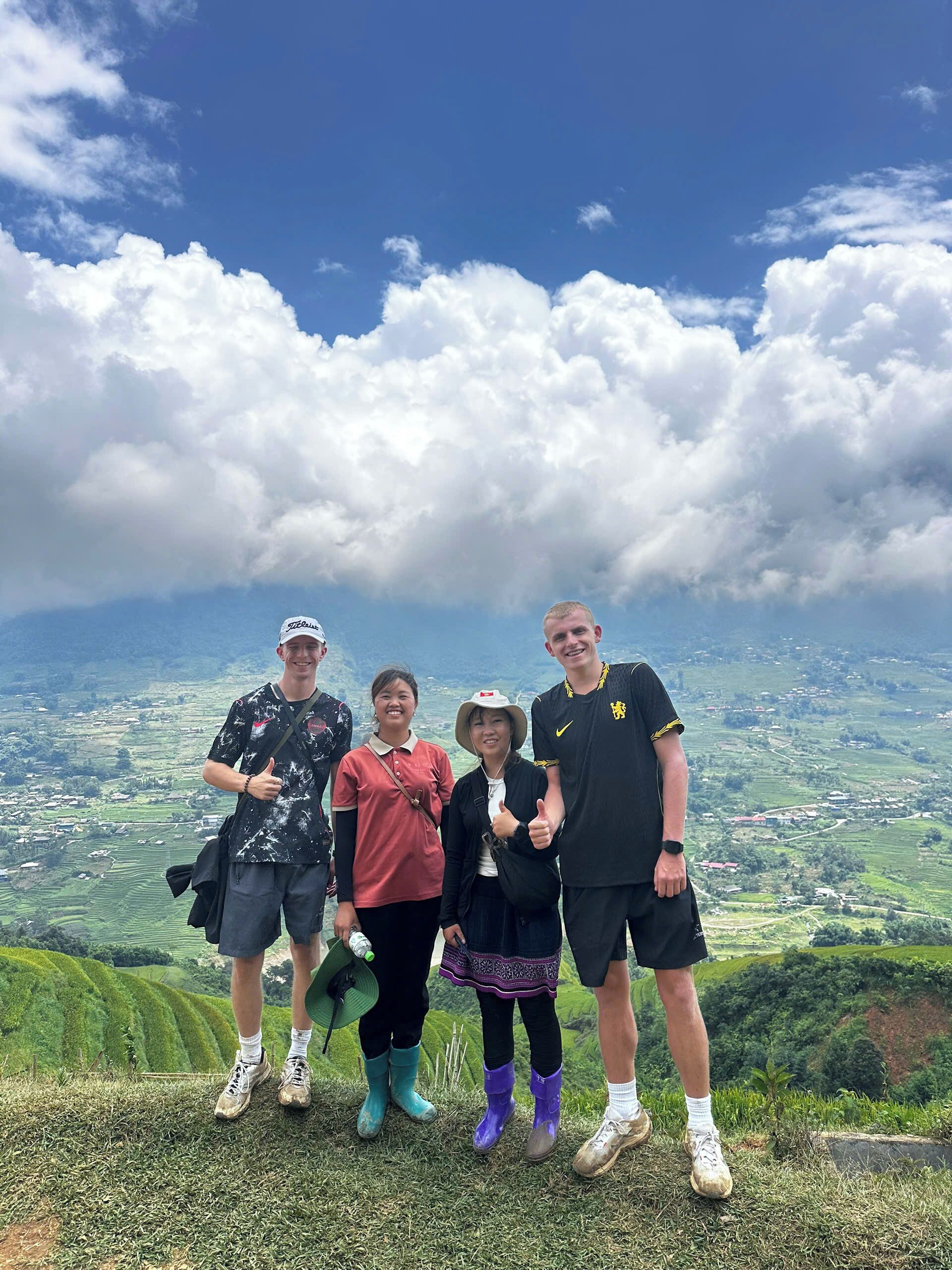 Um grupo de pessoas está em uma colina gramada, apreciando a vista panorâmica de um vale e montanhas ao fundo.