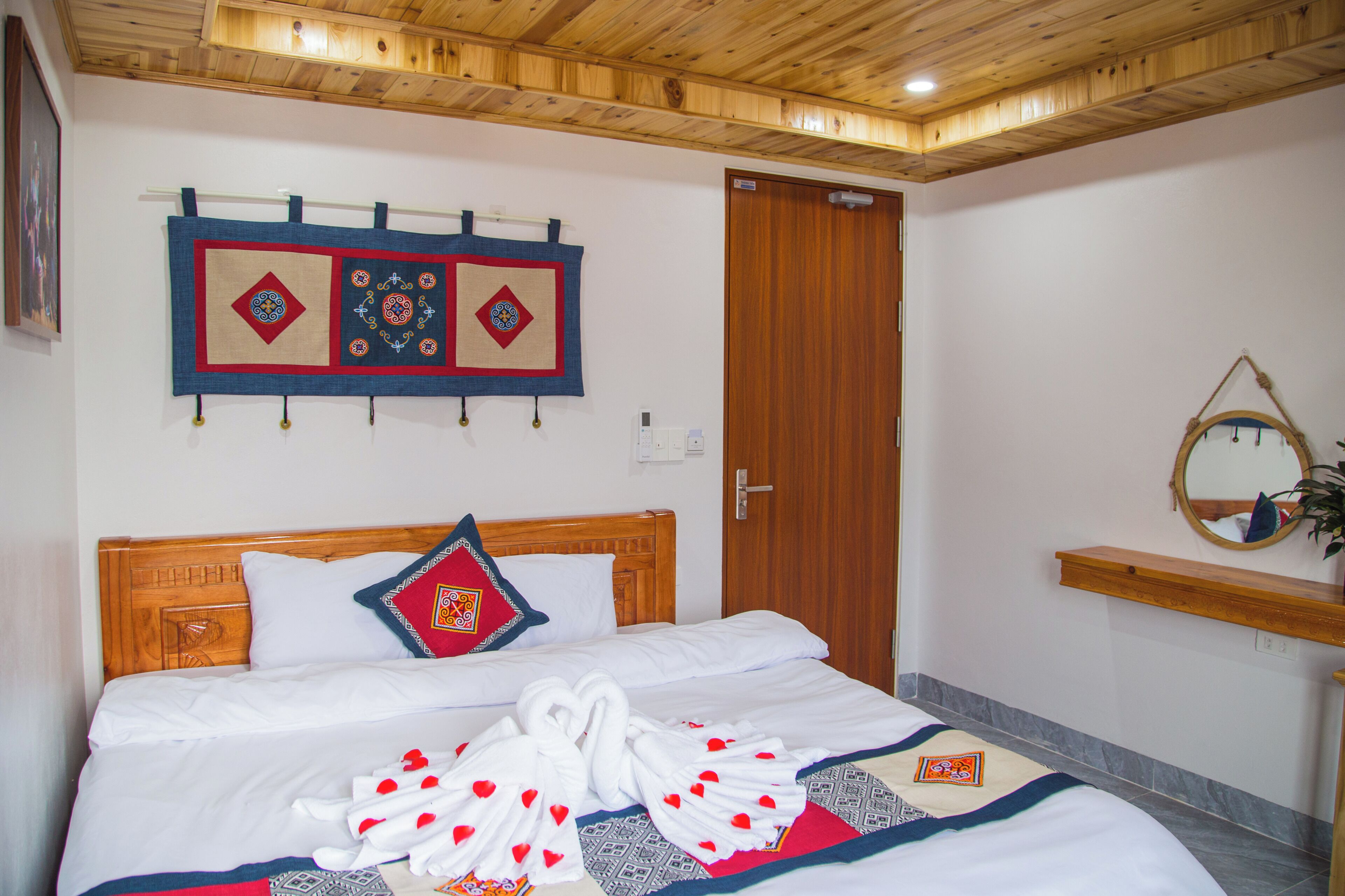 Habitación de hotel con techo de madera, cama con ropa blanca, un tapiz decorativo, puerta de madera y un espejo en la pared.