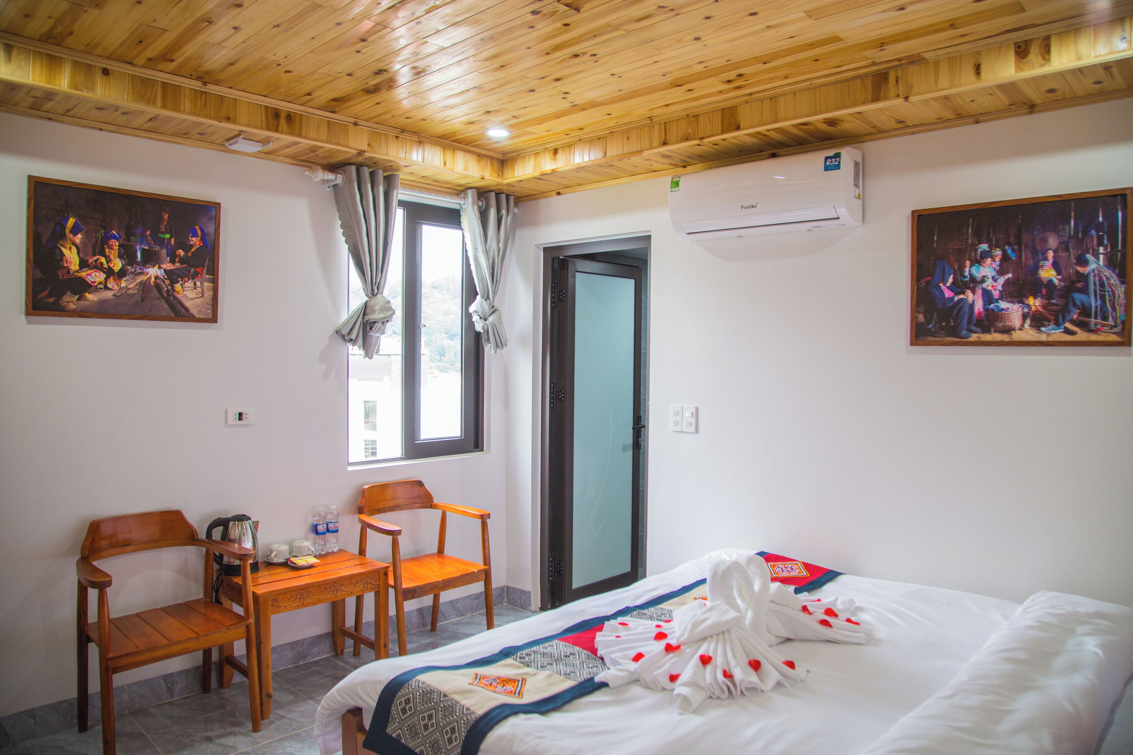 Una habitación de hotel con techo de madera, una cama con cubrecama blanco y decoraciones con forma de corazón, un escritorio de madera con sillas y dos cuadros en las paredes.
