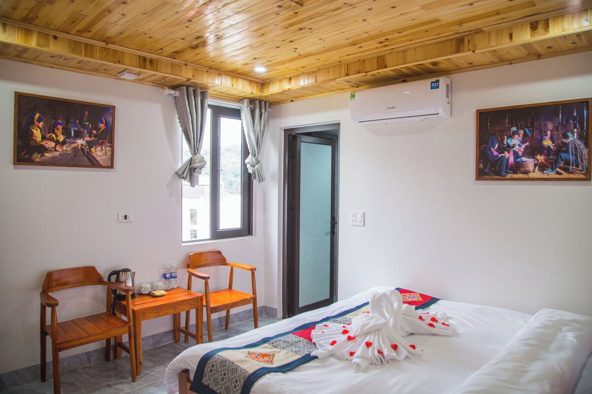 A hotel room with a wooden ceiling, a bed with a white comforter and heart-shaped decorations, a wooden desk with chairs, and two paintings on the walls.
