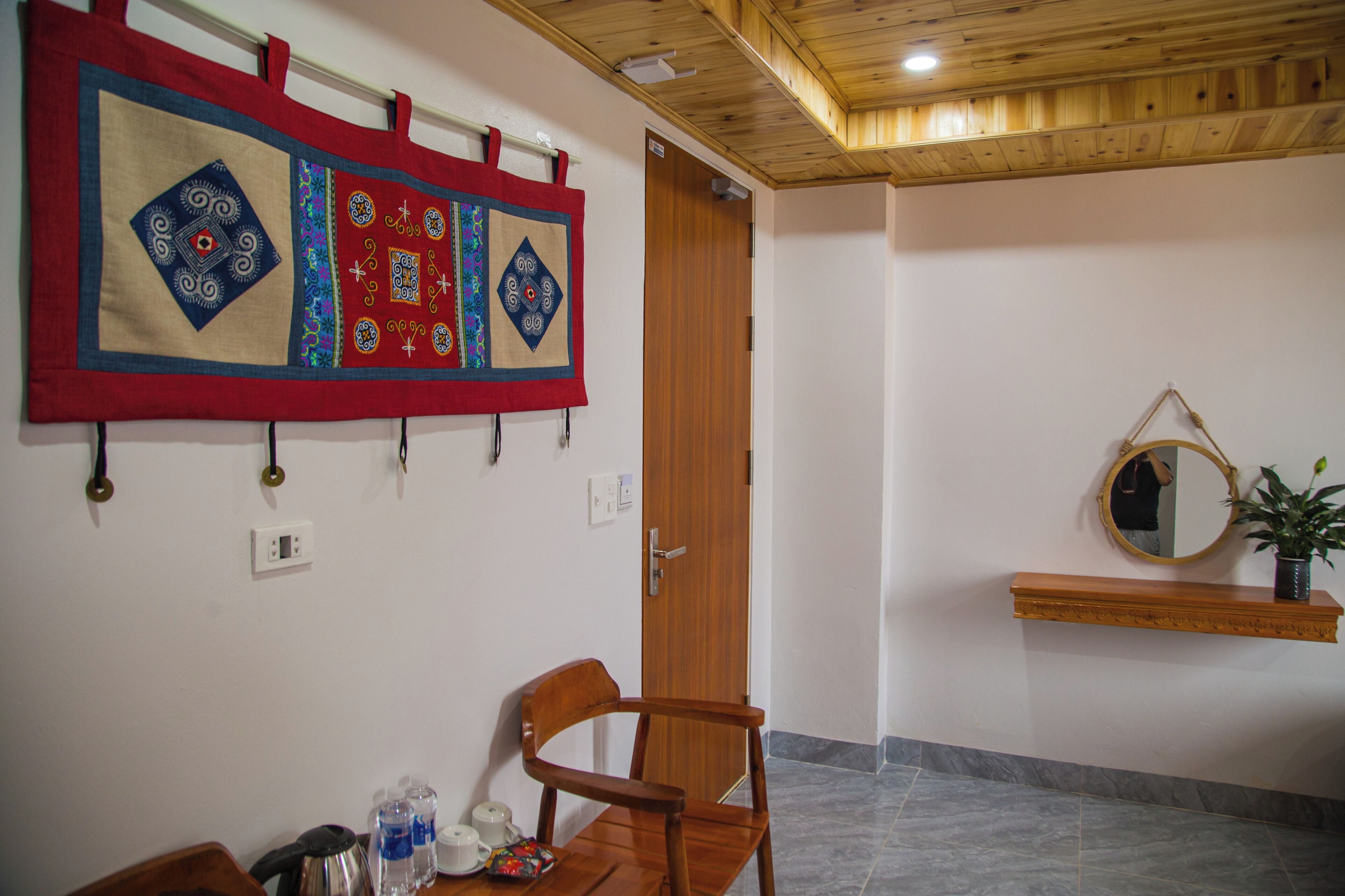 A room with a wooden ceiling, a wooden door, a wall-mounted quilt, a chair, a table with a kettle and water bottles, and a small shelf with a mirror and a plant.