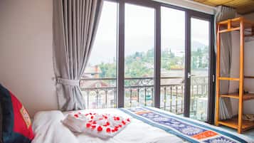 A bedroom with a bed, a window with curtains, a view of buildings, and a wooden shelf.