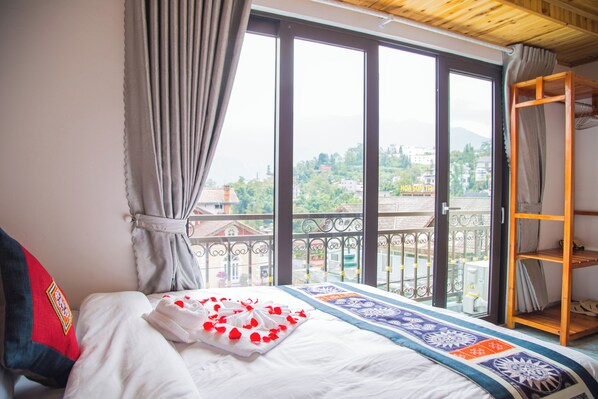 A bedroom with a bed, a window with curtains, a view of buildings, and a wooden shelf.