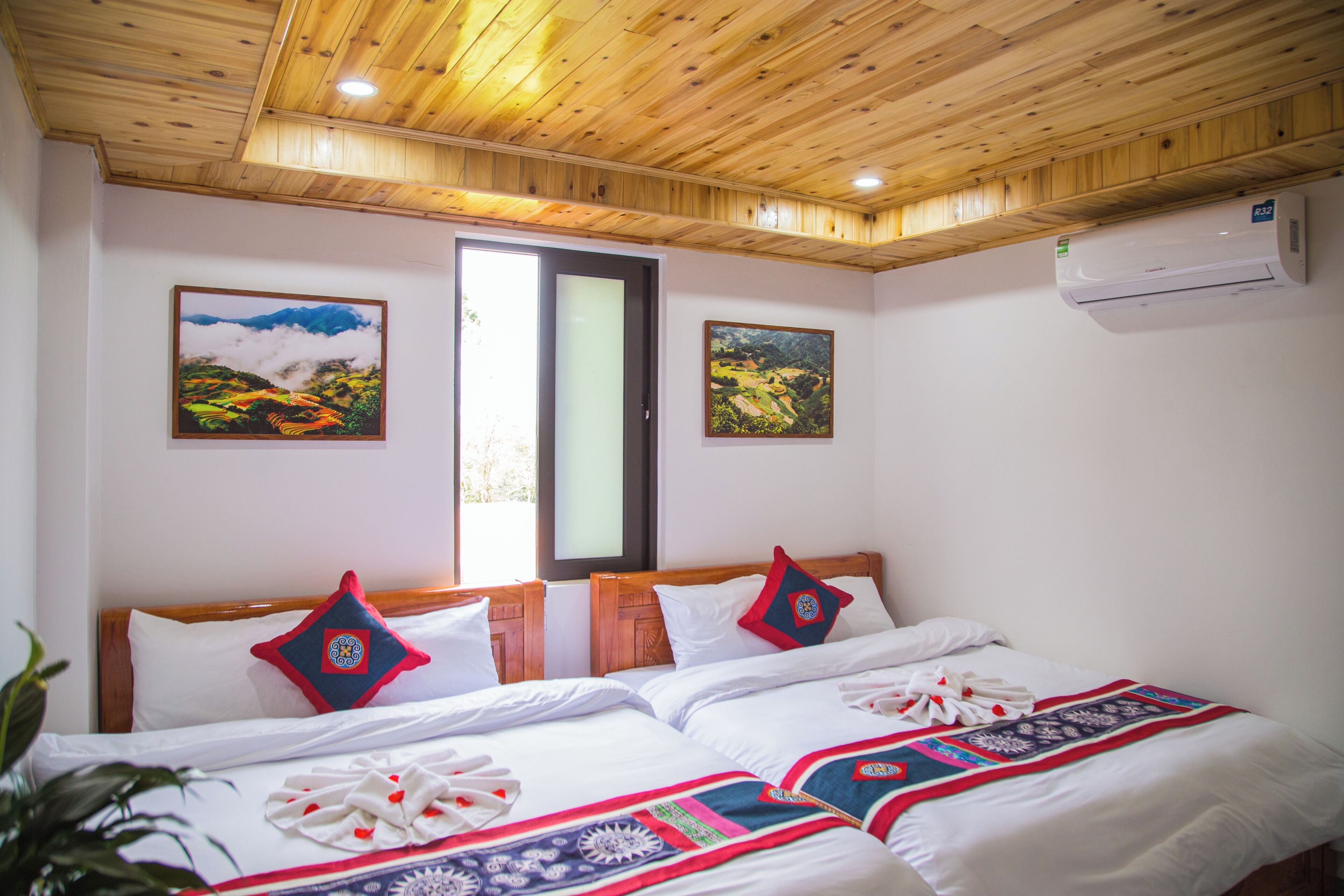 Ein Hotelzimmer mit Holzboden, einem Bett mit weißer Bettwäsche und Dekokissen, zwei Landschaftsbildern an den Wänden sowie einer Klimaanlage.