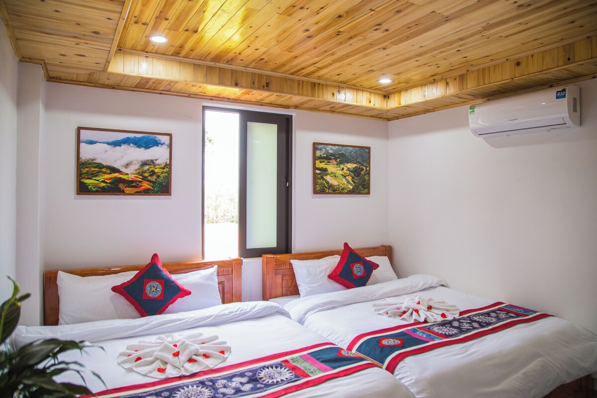 A hotel room with a wooden ceiling, a bed with white bedding and decorative pillows, two landscape paintings on the walls, and an air conditioning unit.
