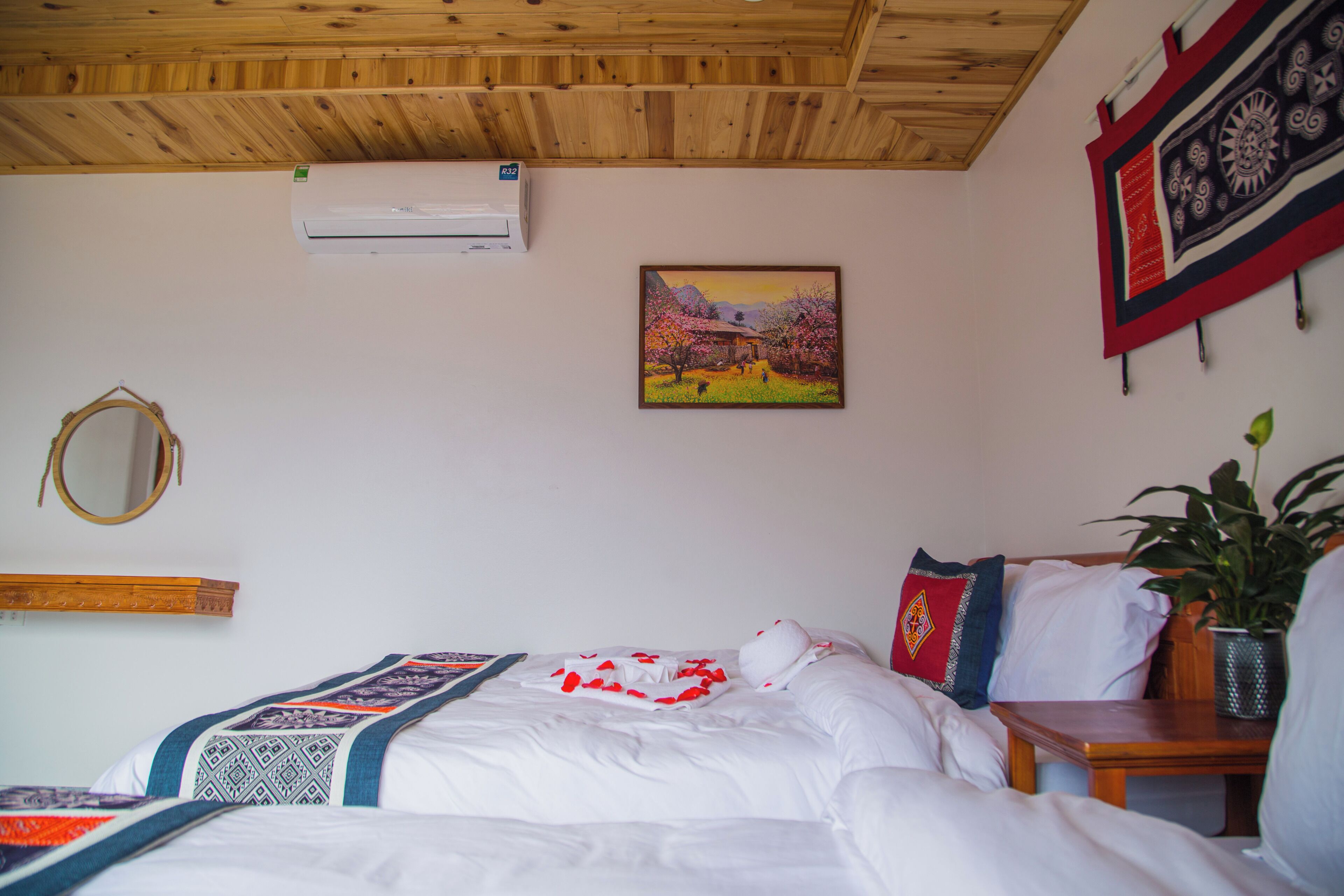 A bedroom with a bed, a bedside table, a potted plant, a wall-mounted air conditioner, a framed painting, and a mirror.